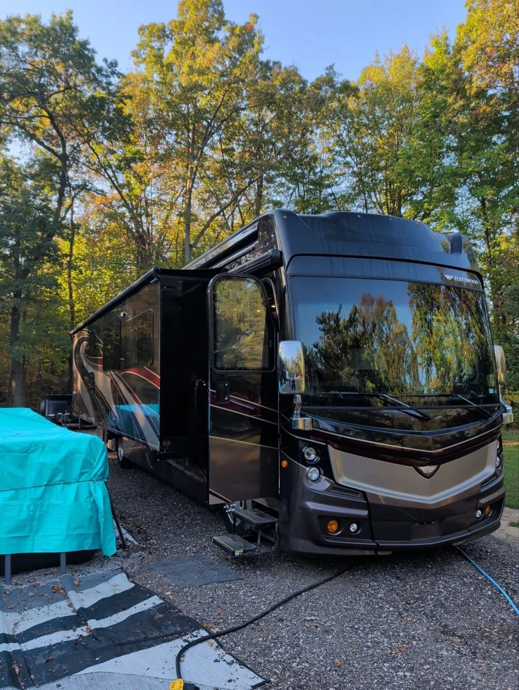 Class A diesel pusher motorhome at a wooded campsite in fall with shore power connected